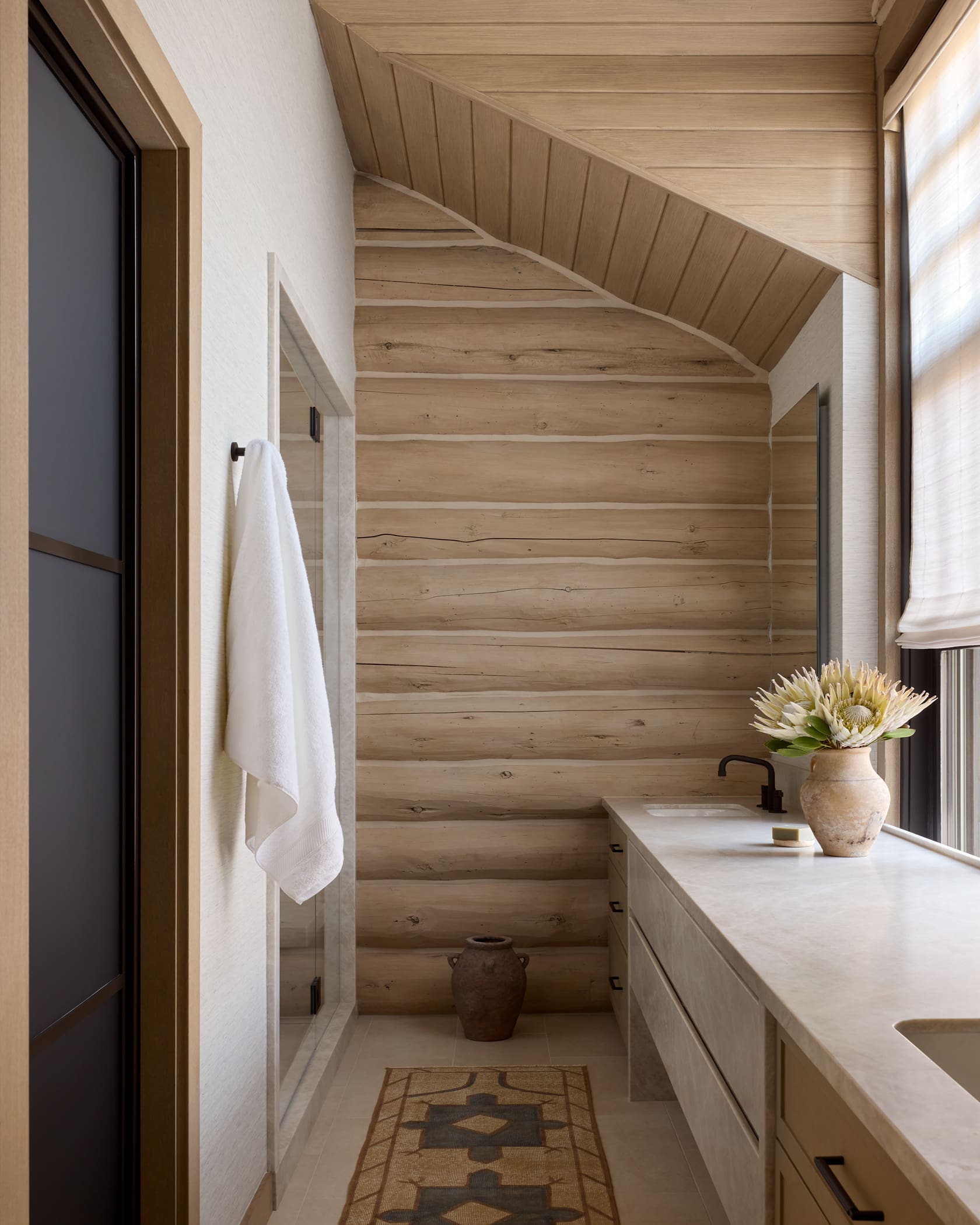 Primary bath with bleached log walls, stone vanity, glass shower, and peaked ceiling, Snowmass log cabin remodel